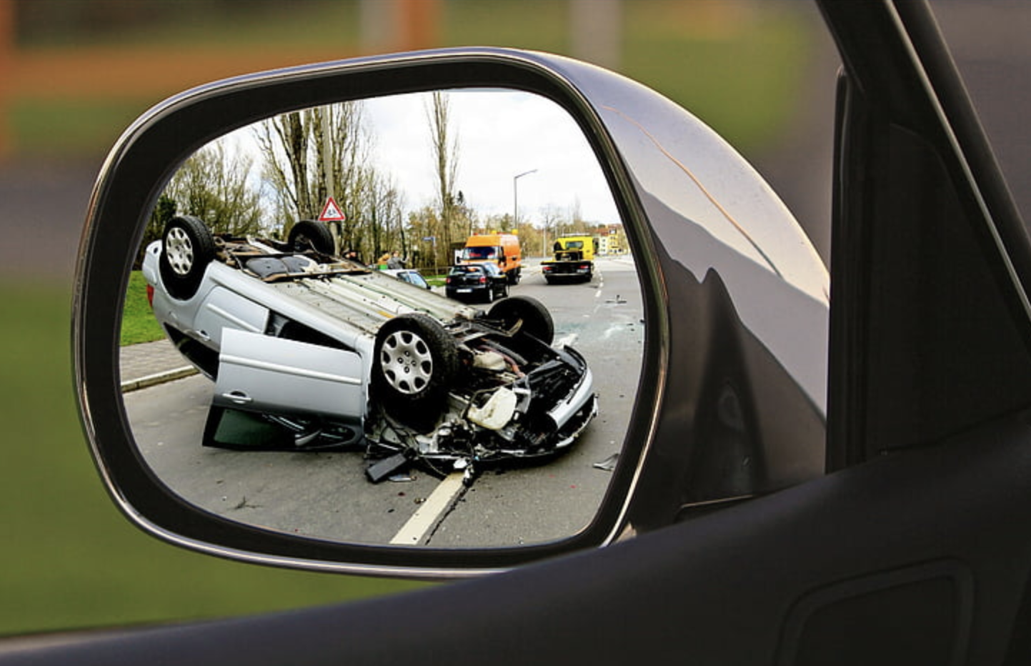 A flipped car from a major accident is visible in the driver’s side mirror, showing severe damage.